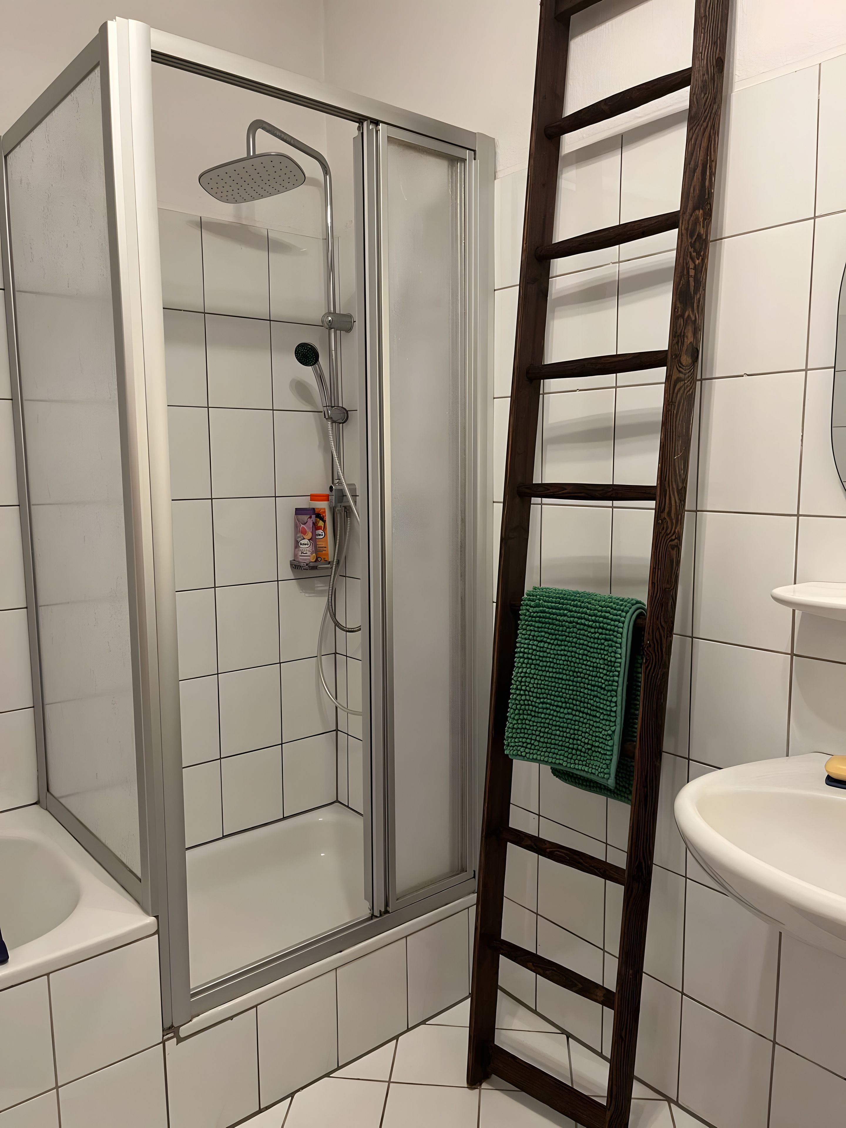 Eine Dusche mit Glastür, eine Holzleiter, ein grünes Handtuch und ein weißes Waschbecken.