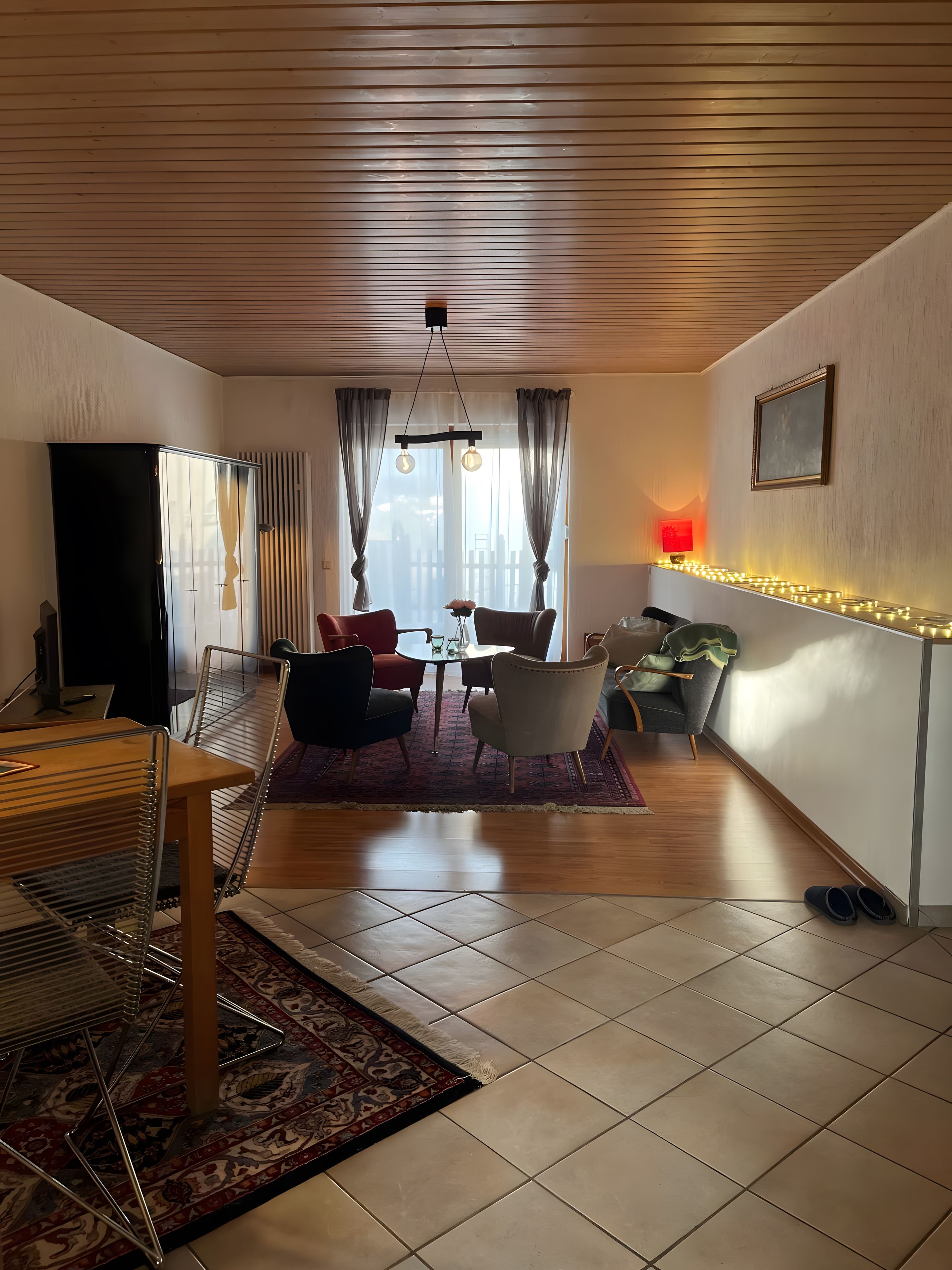 Ein Wohnzimmer mit Holzboden, einem Fernseher, einem Esstisch mit Stühlen, einer Couch, einem gemusterten Teppich und einer Wandleuchte.