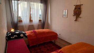 A small, cozy room with two beds, a window with sheer curtains, and a wall-mounted decorative item.