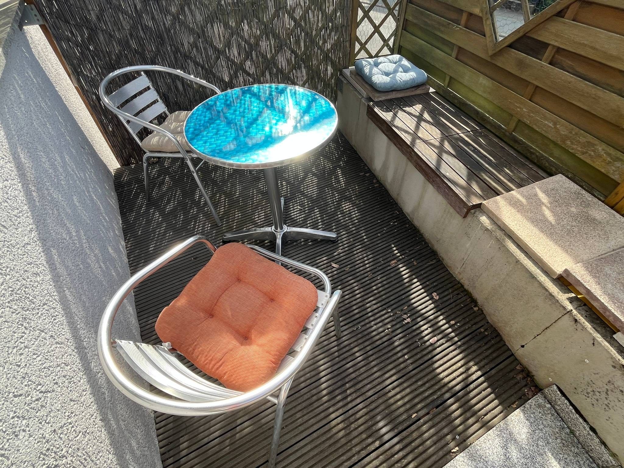 A patio area with a round table and two chairs, a small round table with a cushion, and a wooden bench with a cushion.