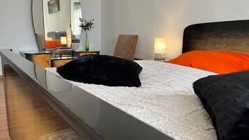 Een slaapkamer met een bed, nachtkastje, lamp, spiegel en een vaas met bloemen.