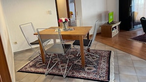 Un coin repas avec une table en bois, des chaises en métal et un tapis à motifs. Un téléviseur est fixé au mur, et on peut voir un porte-chaussures.