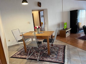 Ein Essbereich mit einem Holztisch, Metallstühlen und einem gemusterten Teppich. Ein Fernseher ist an der Wand montiert, und ein Schuhregal ist zu sehen.
