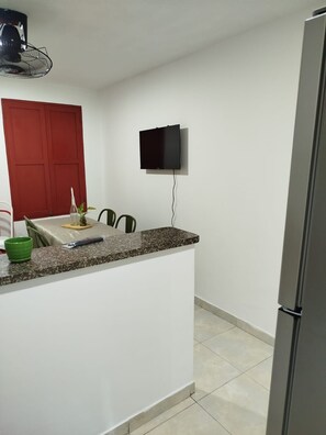 Eine kompakte Küche mit Granit Arbeitsplatte, einem an der Wand befestigten Flachbildfernseher, einem Kühlschrank und einem Essbereich mit roter Tür und Tisch.