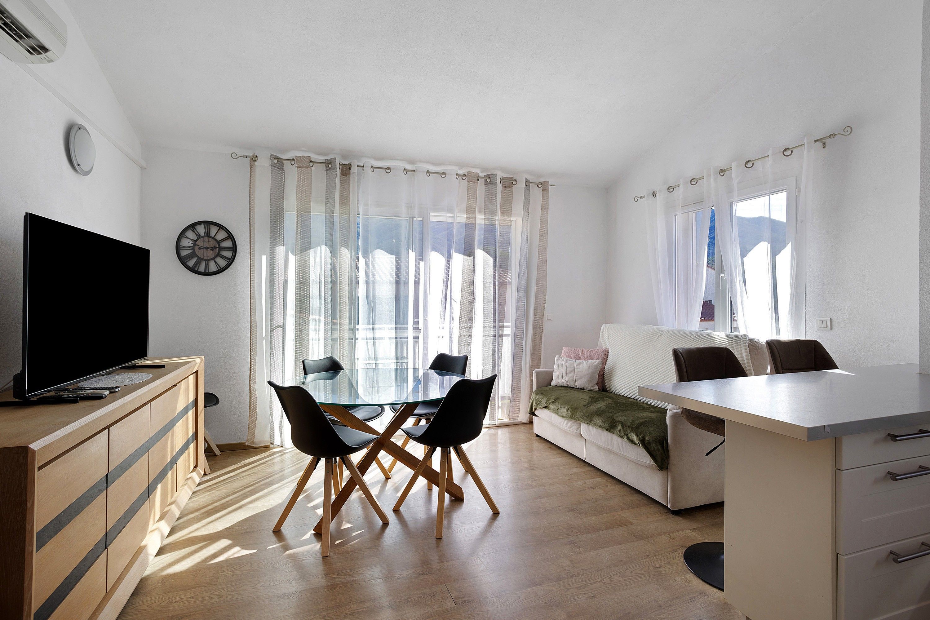 Een moderne woonkamer met een flatscreen televisie, een eettafel met stoelen, een bank en een groot raam met lichte gordijnen.