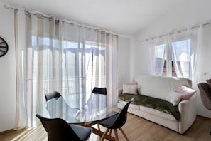 A modern living room with a glass dining table, black chairs, a white sofa with a green throw, and sheer curtains.