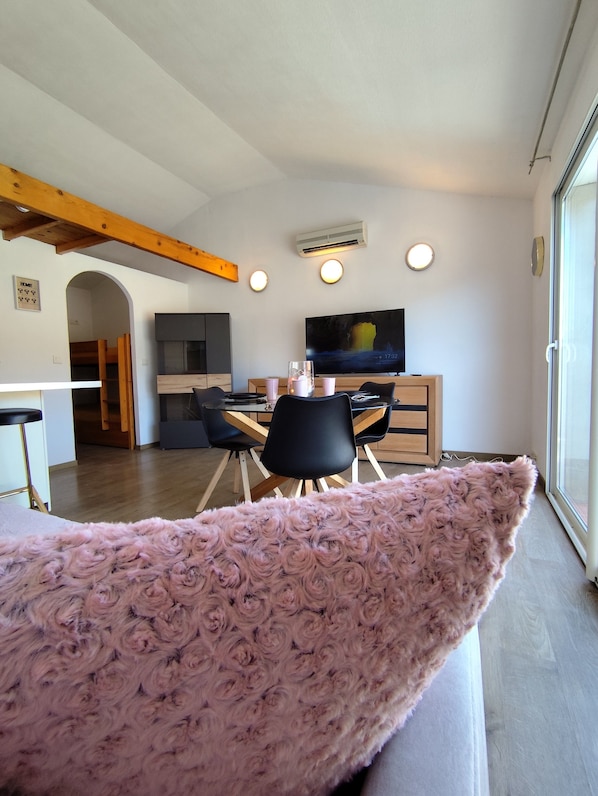 Een moderne woonkamer met een groot raam, een flatscreen televisie, een eethoek met stoelen en een zacht roze vloerkleed.