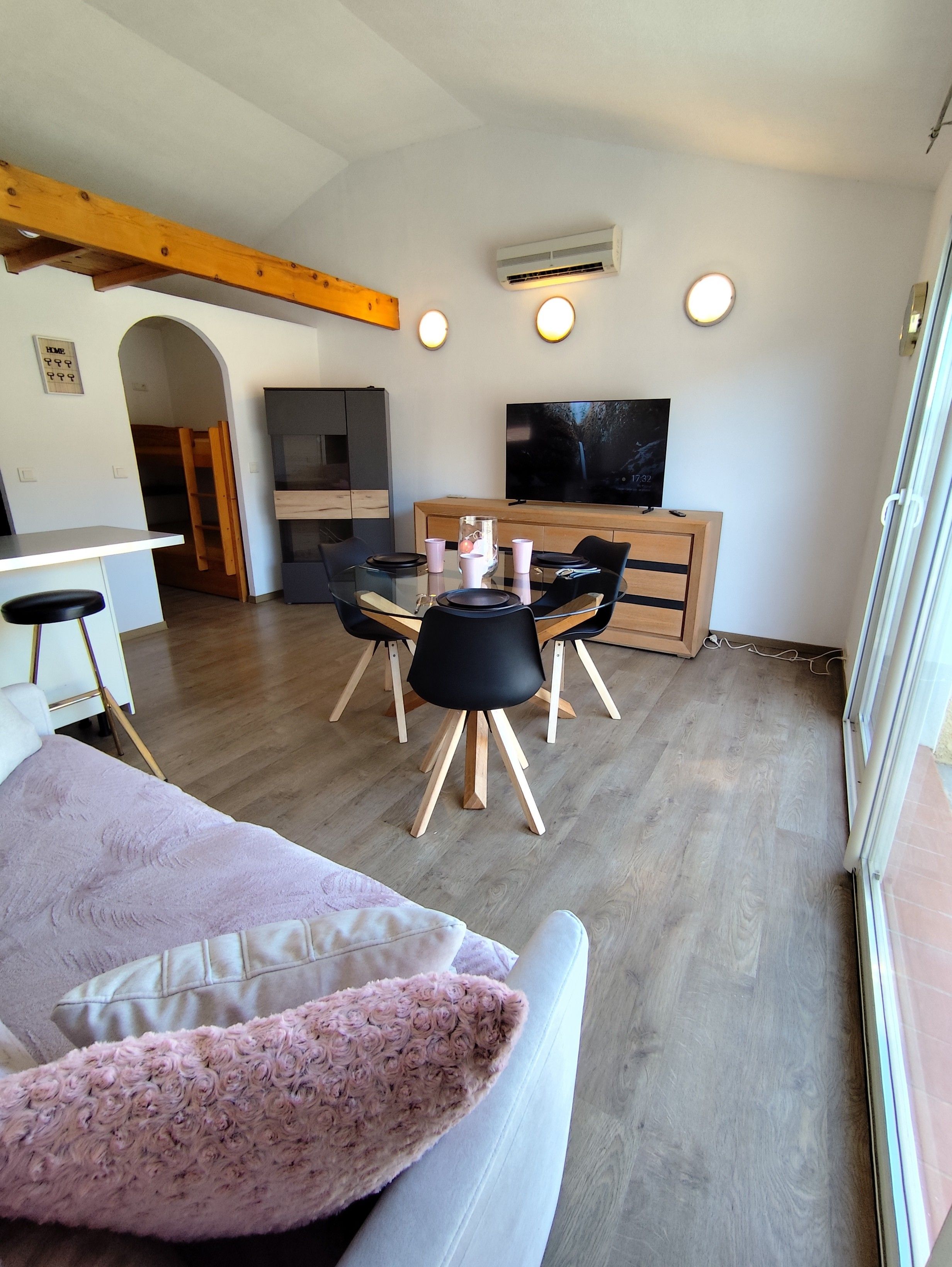 A modern living room with a dining table, chairs, a flat-screen TV, and a wooden ceiling beam.
