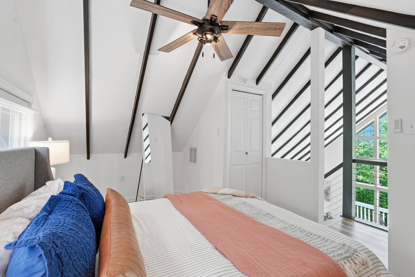 Een slaapkamer met schuine wanden, een plafondventilator, een bed met veel kussens, een nachtkastje en een raam met uitzicht op groen.