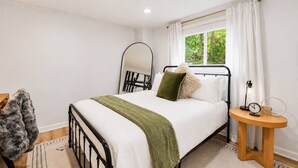 Een slaapkamer met een bed, nachtkastje, spiegel en raam met gordijnen.