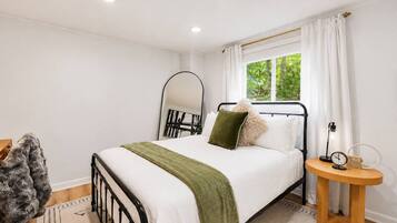 Quarto com cama, mesa de cabeceira, espelho e janela com cortinas.