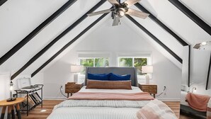 Een slaapkamer met een schuine kap, een bed met gestreepte lakens, twee nachtkastjes met lampen en een raam met uitzicht op groen.