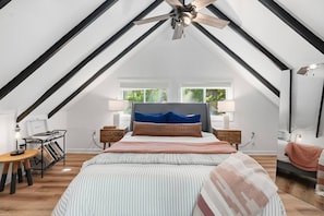 A bedroom with a sloped ceiling, a bed with striped bedding, two bedside tables with lamps, and a window with a view of greenery. - A-Frame Soak and Cedar near Skyline Drive and Wineries (Front Royal)