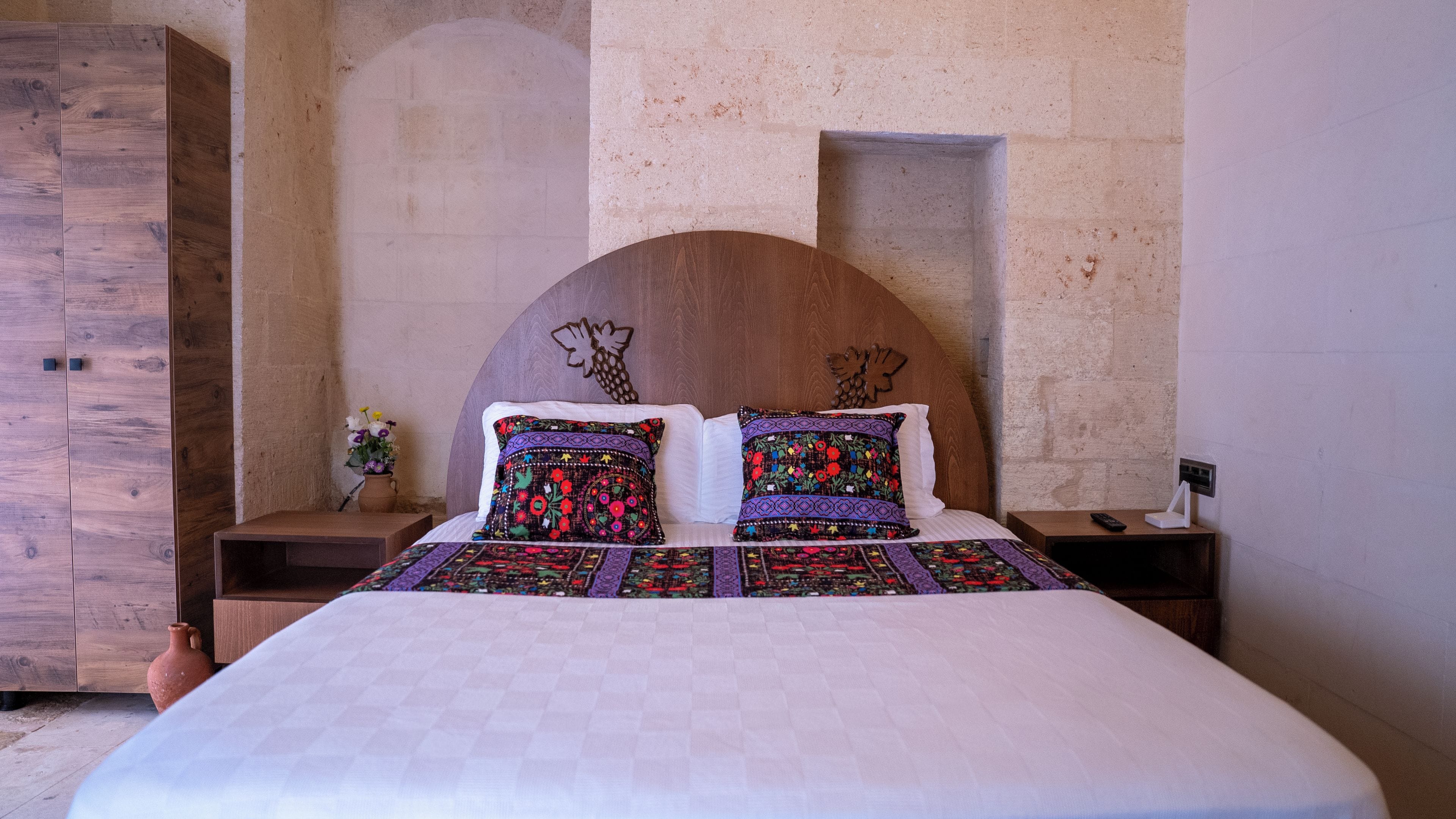 Una cama con cabecera de madera, dos almohadas con motivos florales, una mesita de noche con un control remoto y un florero con flores.