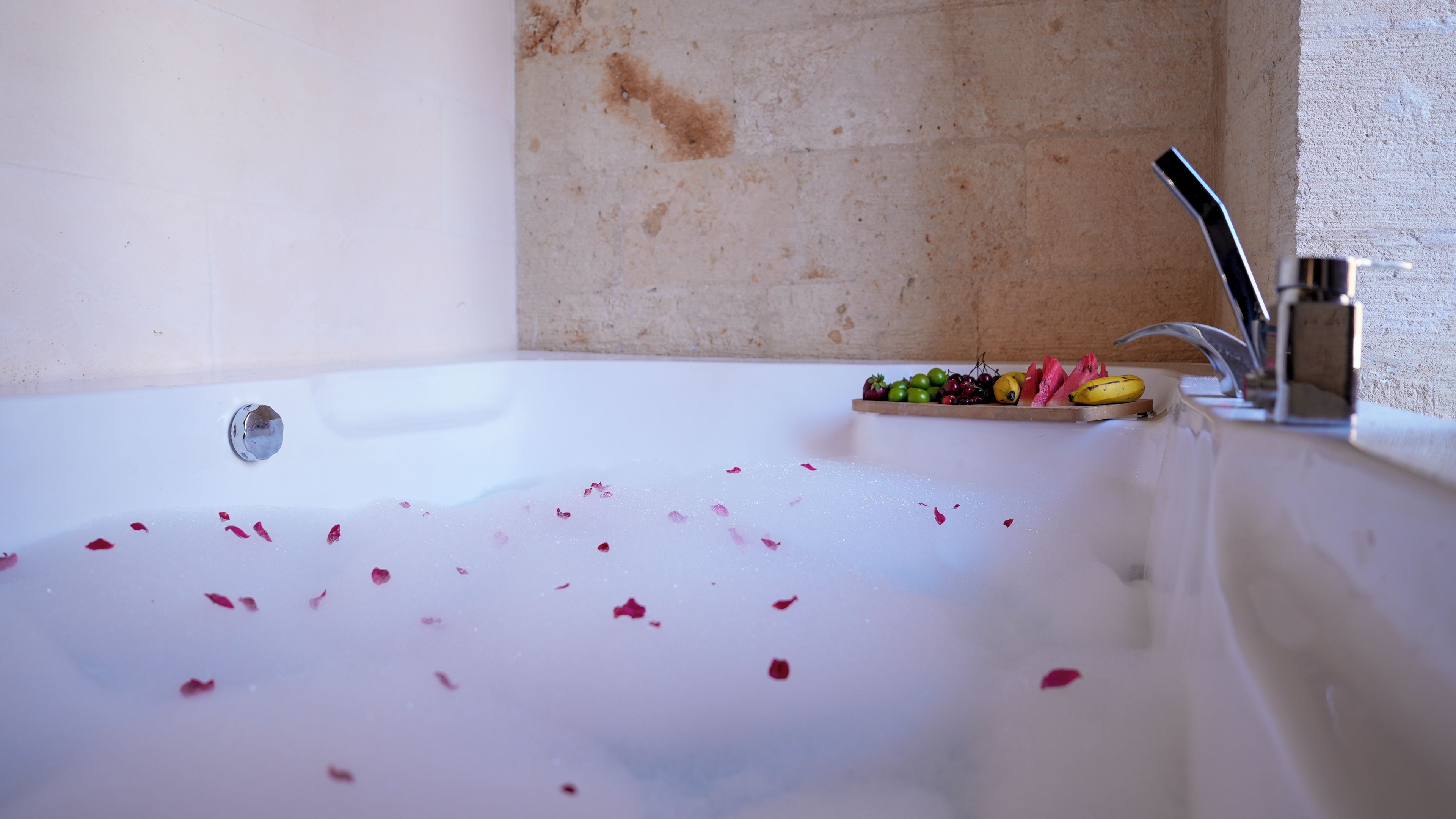 Eine Badewanne mit darin treibenden Rosenblüten, eine Obstschale am Rand und ein moderner Wasserhahn.