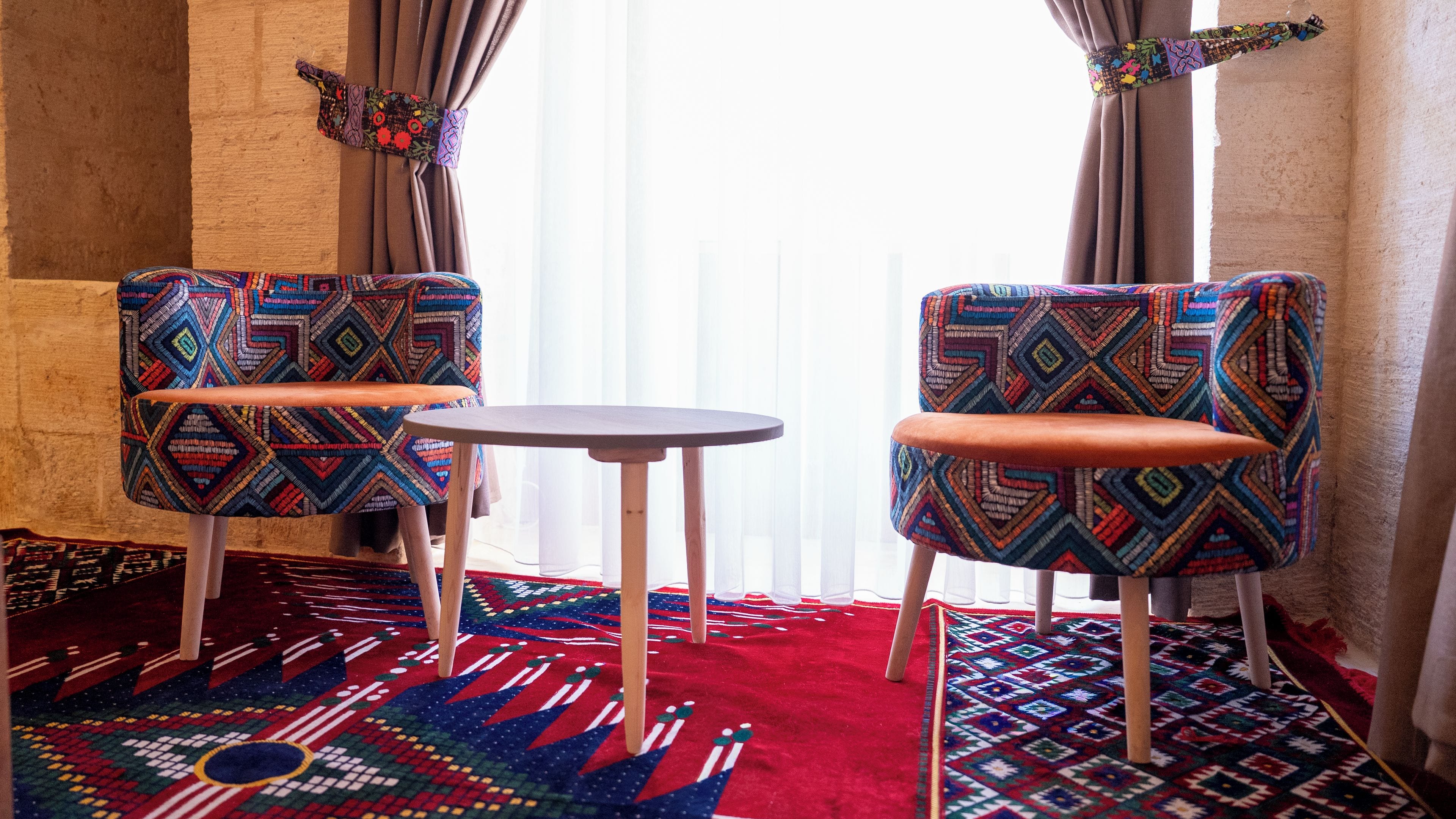 Two chairs with patterned upholstery, a small round table, and a red patterned rug in a room with a wooden floor and a window with curtains.