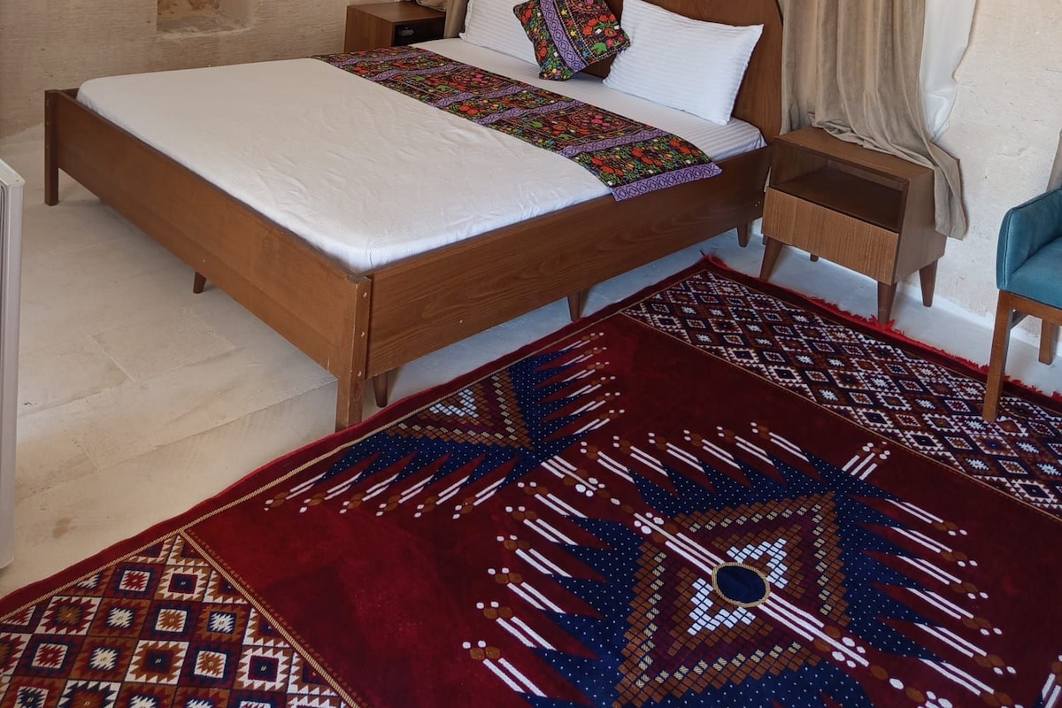 Un dormitorio con cama de madera, mesita de noche, alfombra estampada y ventana con cortinas.