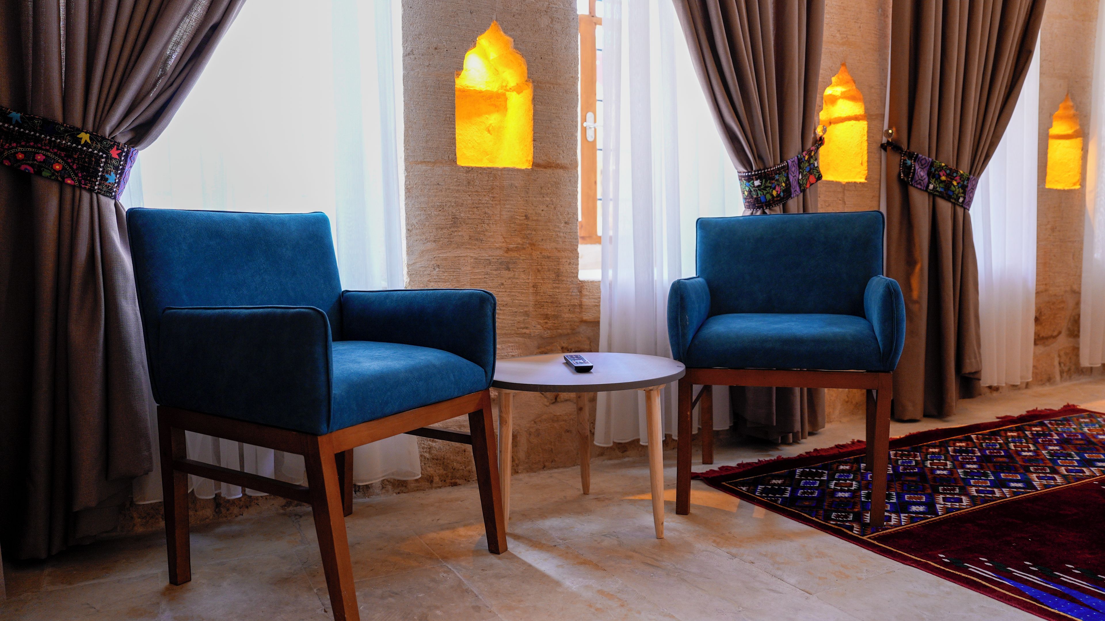 Two blue armchairs, a small round table, and a patterned rug in a room with a brick wall and decorative lighting.