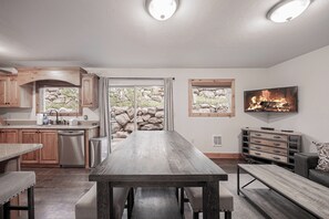 A modern kitchen with a stone wall backdrop, a dining area with a long table, and a fireplace with a TV. - Walk to Ski Lifts | 2 Living Areas | Sleeps 8 (Brian Head)