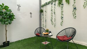 Una piccola area salotto all'aperto con due sedie e un tavolo, circondata da erba sintetica e un muro bianco con piante pensili.