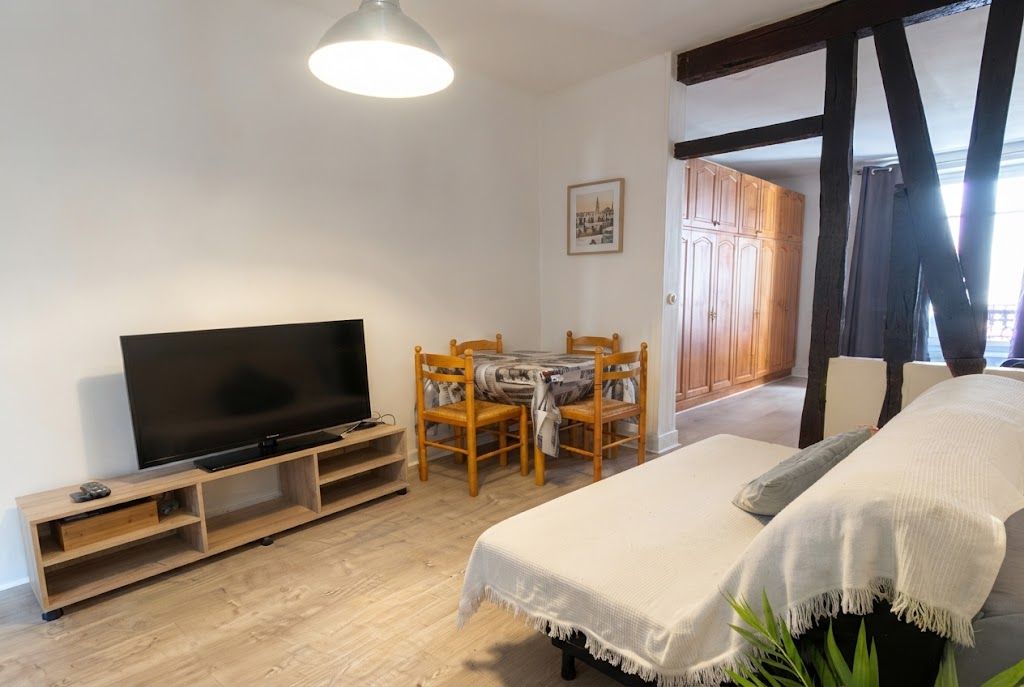 Una habitación con televisión sobre un mueble de madera, una mesa de comedor con sillas, una cama con edredón blanco y un armario de madera.