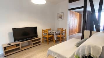 Una habitación con televisión sobre un mueble de madera, una mesa de comedor con sillas, una cama con edredón blanco y un armario de madera.