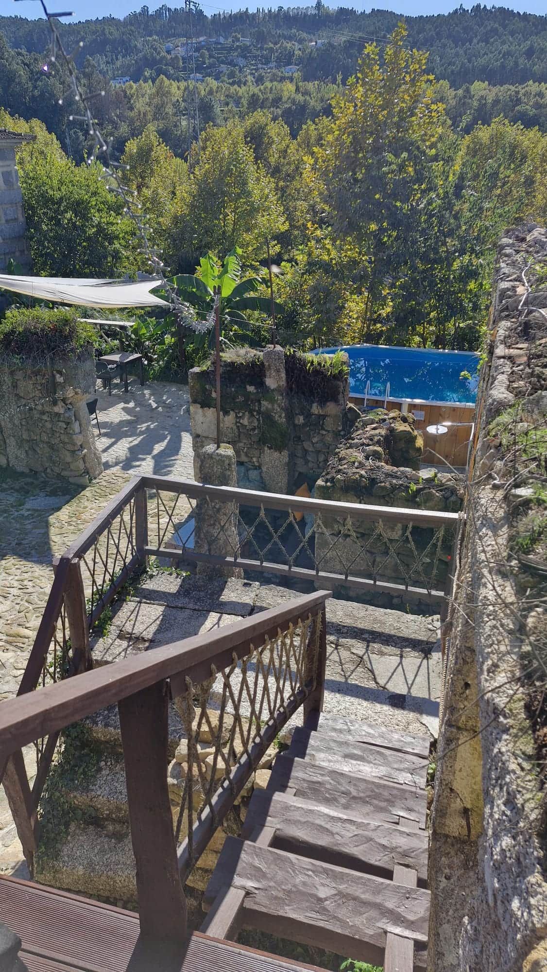 Un escalier en bois mène à une plateforme en pierre, bordée d’une rambarde en métal, et est entouré de verdure. On peut admirer une vue sur une colline depuis cet endroit.