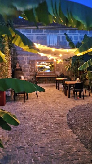 Un patio al aire libre con suelo de piedra, una mesa de madera y sillas, rodeado de follaje e iluminado con luces colgantes.
