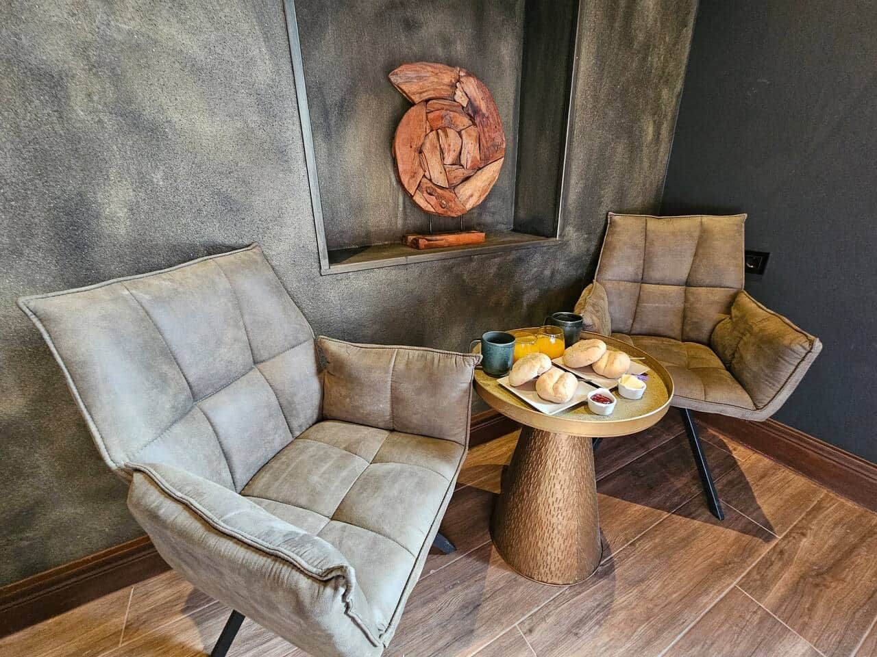 Ein Zimmer mit zwei Polstersesseln, einem runden Holztisch mit Frühstücksgebäck und einer an der Wand befestigten Holzskulptur.
