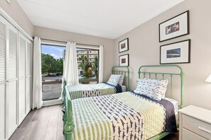 A bedroom with two beds, a window with curtains, a green metal headboard, and a small bedside table. - Litchfield By The Ocean, Two bedroom condo across from Litchfield Beach (Pawleys Island)