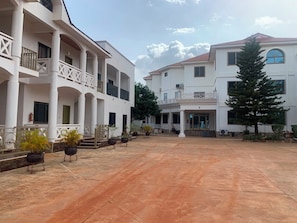 A spacious courtyard with a covered walkway, potted plants, and a building with multiple balconies and a central entrance.