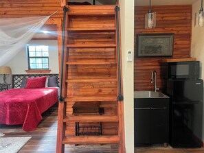 Eine Holztreppe führt zu einem Schlafzimmer auf dem Dachboden, ausgestattet mit einer roten Bettdecke, einer kleinen Küche mit einem schwarzen Kühlschrank und einer hängenden Leuchte.
