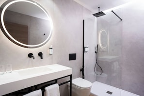 Un baño moderno con un espejo redondo, un lavamanos blanco, un inodoro y una ducha con mampara de cristal.