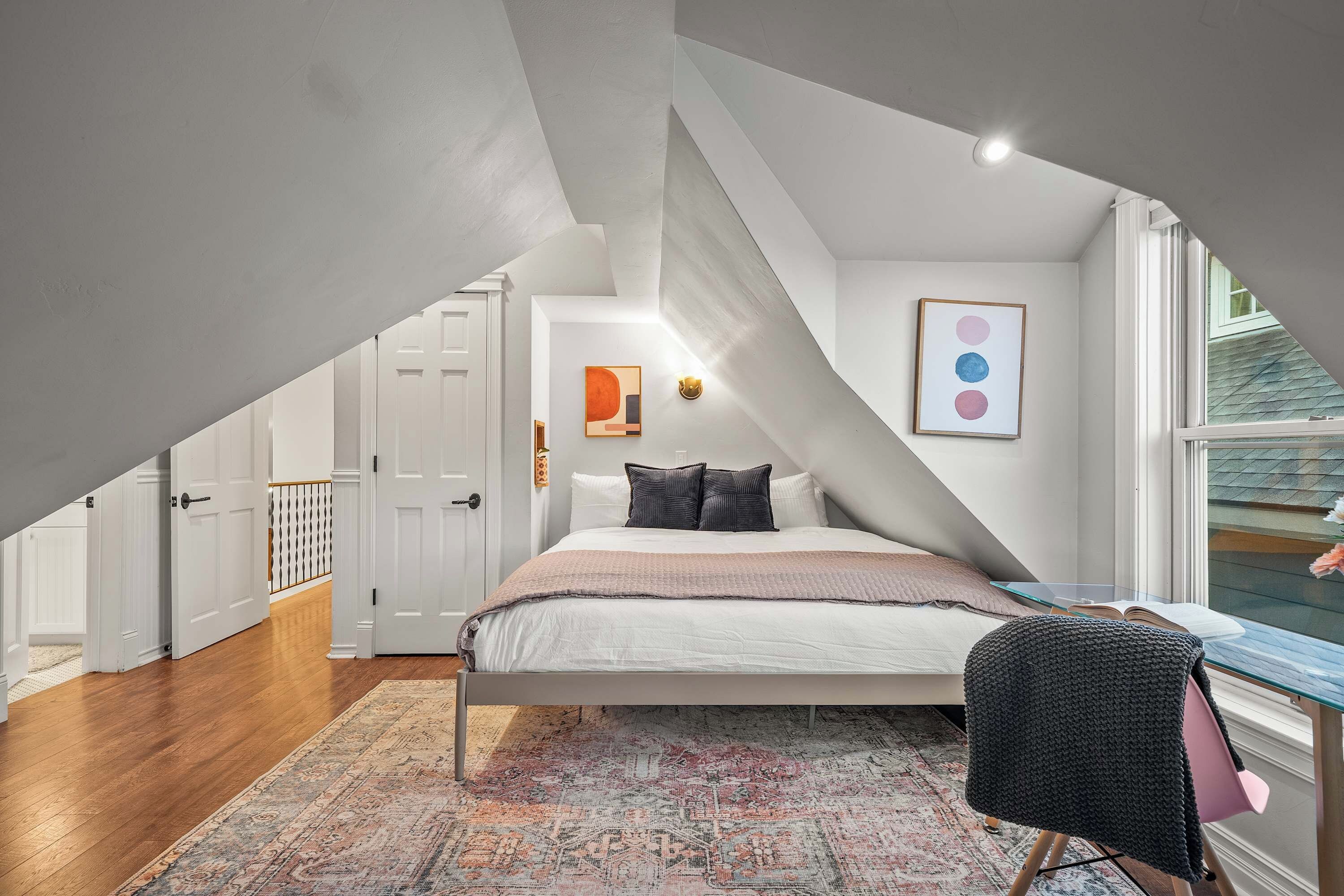 Une chambre avec un plafond en pente, un lit avec des oreillers, un tapis, une chaise et des œuvres d’art accrochées au mur.