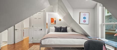 A bedroom with a sloped ceiling, a bed with pillows, a rug, a chair, and artwork on the wall.