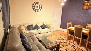 Ein Wohnzimmer mit einer Couch, einem Couchtisch, einem Esstisch mit Stühlen, einer Wanduhr und einer Tür.