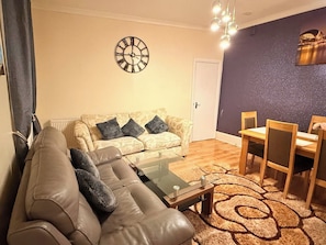 A living room with a sofa, a coffee table, a dining table with chairs, a clock on the wall, and a door. - Spacious 4BR House | 5 Mins to city | Sleeps 9 (Tyne and Wear)