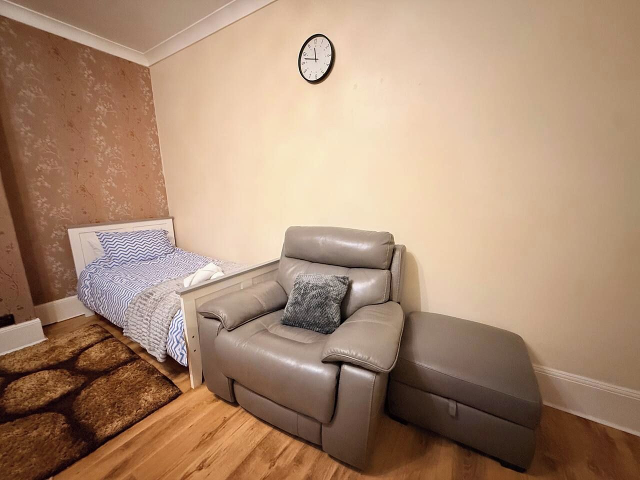 Une chambre à coucher avec un lit, un fauteuil de massage gris, une horloge au mur et un tapis à motifs.