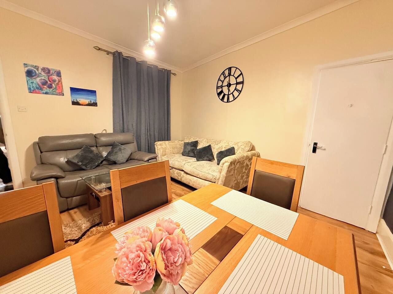 Una sala de estar con un sofá, una mesa de comedor con sillas, un reloj en la pared y una puerta.