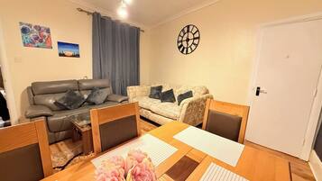 Una sala de estar con un sofá, una mesa de comedor con sillas, un reloj en la pared y una puerta.