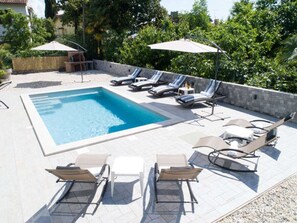 Ein Schwimmbecken mit Liegestühlen und Sonnenschirmen, umgeben von einer Steinmauer und viel Grün.