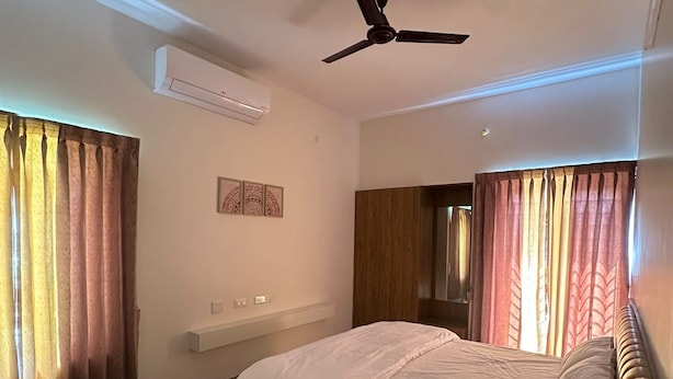 A hotel room with a bed, a ceiling fan, an air conditioning unit, a vanity with a mirror, and curtains. - MV Home Stay (Thirumangalam)
