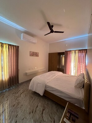 A hotel room with a bed, a ceiling fan, an air conditioning unit, a vanity with a mirror, and curtains.