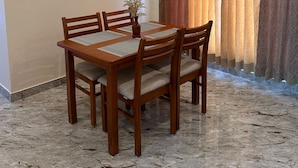 A dining area with a wooden table and chairs, a vase with dried flowers, and a curtain with a patterned design. - MV Home Stay (Thirumangalam)