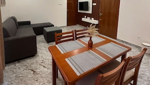A modern living room with a dining area, a sofa set, a TV mounted on the wall, and ceiling fans. - MV Home Stay (Thirumangalam)