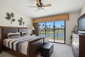 A bedroom with a bed, a nightstand, a lamp, a TV, a dresser, a ceiling fan, and a large window with a view of a golf course and palm trees. - Palm Valley Oasis with Golf Course & Lake Views (Palm Desert)