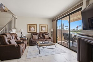 A spacious living room with a brown leather sofa, a glass coffee table, and a large sliding glass door leading to an outdoor patio. - Palm Valley Oasis with Golf Course & Lake Views (Palm Desert)