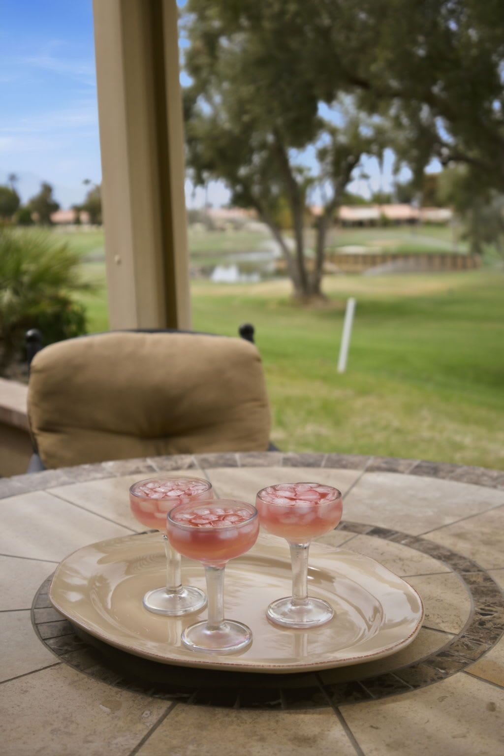 A patio table with a glass tray holding three glasses of pink beverage, a beige chair, and a view of a golf course.