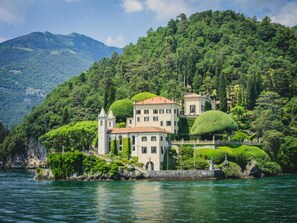 A lakeside property with a large building featuring multiple windows and a tower, surrounded by lush greenery and hills. - Apartment in Acquaseria With Terrace (San Siro)