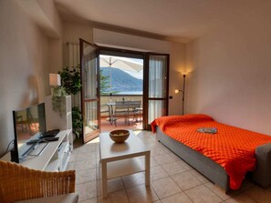 A hotel room with a bed, a desk with a computer, a chair, a table, and a balcony with a view of the mountains. - Apartment in Acquaseria With Terrace (San Siro)